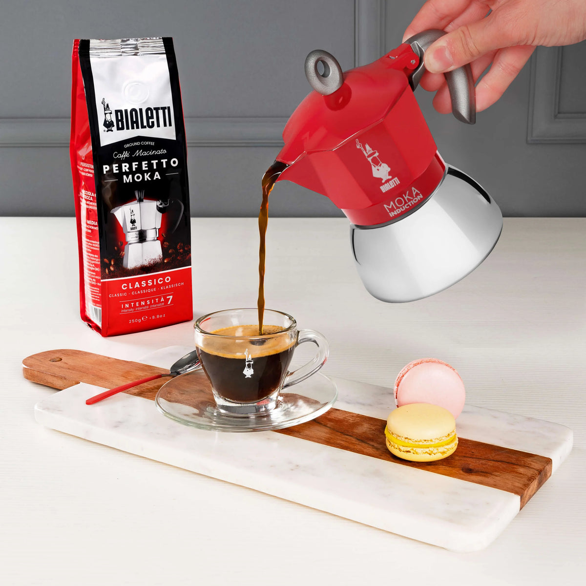 Bialetti coffee maker pouring freshly brewed coffee into a glass cup with macaroons on a wooden board