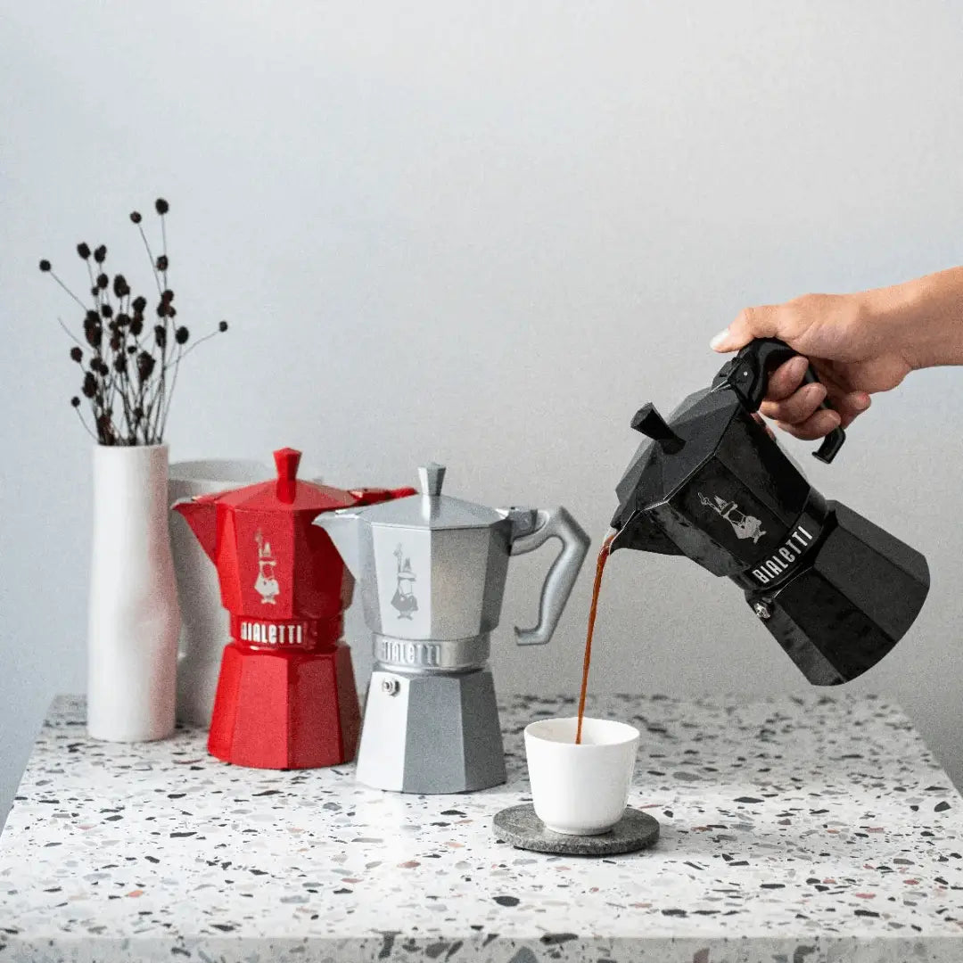 Bialetti Moka Express coffee maker pouring coffee into a cup with red and grey models in the background