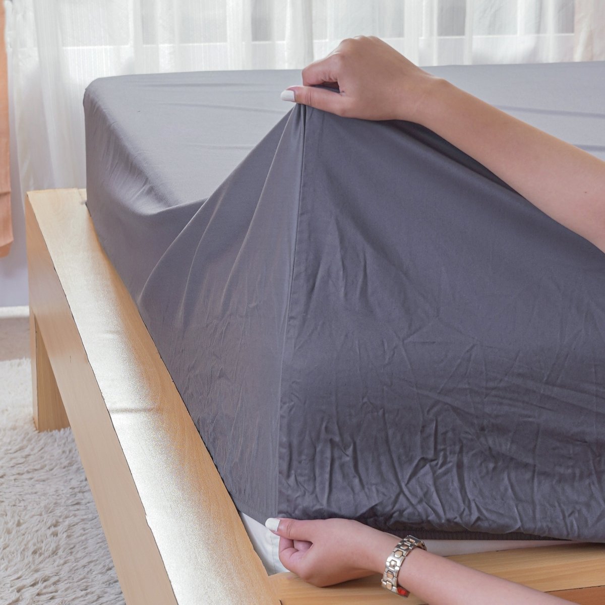 Person putting on a grey fitted sheet from SHEET STORY on a bed with wooden frame