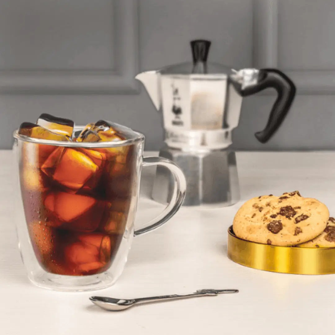 Bialetti glass mugs filled with iced coffee and ice cubes, alongside cookies and a coffee maker