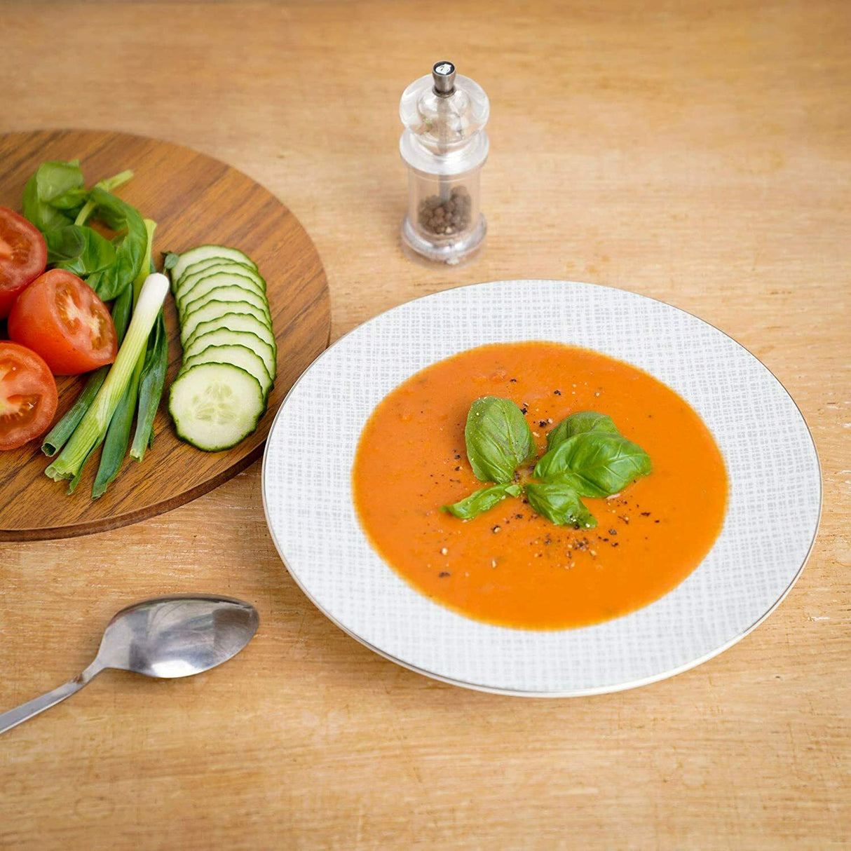 Elegant white soup bowl filled with tomato soup, garnished with basil, on a wooden table with fresh vegetables