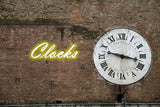 Orsina Wall Clock on a brick wall with a neon sign reading Clocks in the background