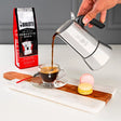 Bialetti Coffee Maker pouring freshly brewed coffee into a glass cup with macarons on a wooden tray