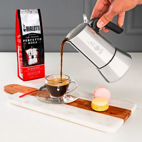 Bialetti Coffee Maker pouring freshly brewed coffee into a glass cup with macarons on a wooden tray