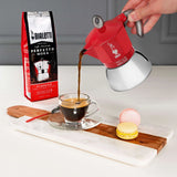 Bialetti coffee maker pouring freshly brewed coffee into a glass cup with macaroons on a wooden board