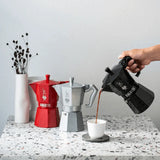 Bialetti Moka Express coffee maker pouring coffee into a cup with red and grey models in the background