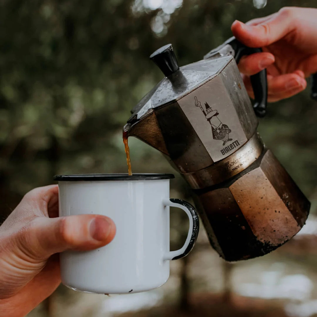Bialetti Moka Express coffee maker pouring fresh coffee into a white cup outdoors