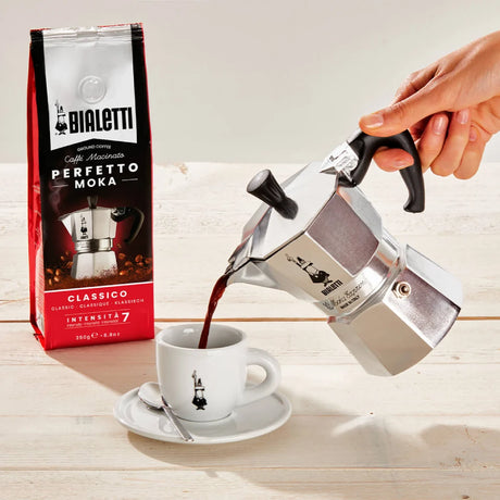 Bialetti Moka Express coffee maker pouring rich coffee into a cup with packaging in the background