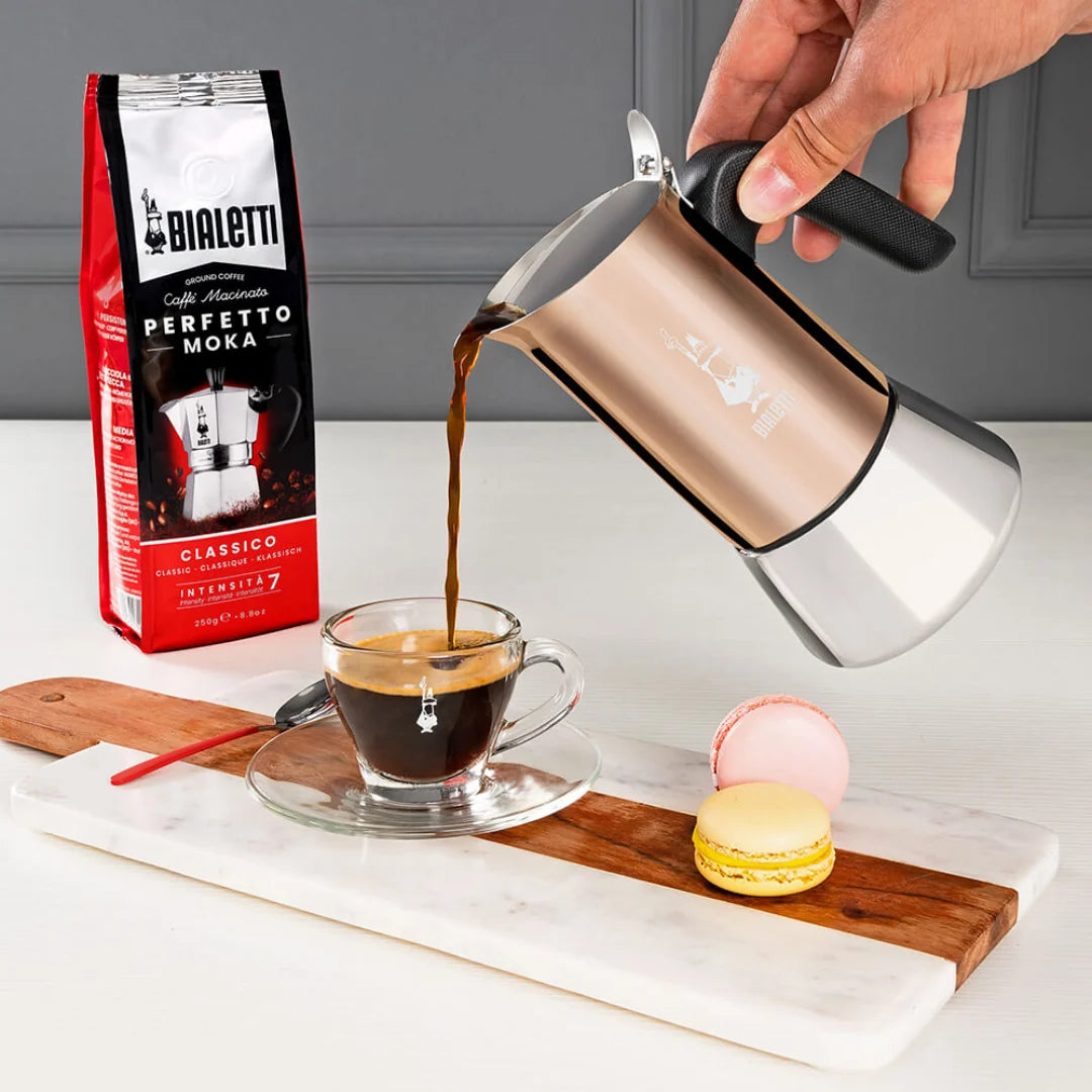 Bialetti coffee maker pouring fresh coffee into a glass cup on a wooden board with macarons