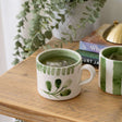 Hand-painted Selbrae House ceramic mug with green accents on a wooden table next to books and a plant