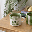 Hand-painted Selbrae House ceramic mug with green accents on a wooden table next to books and a plant