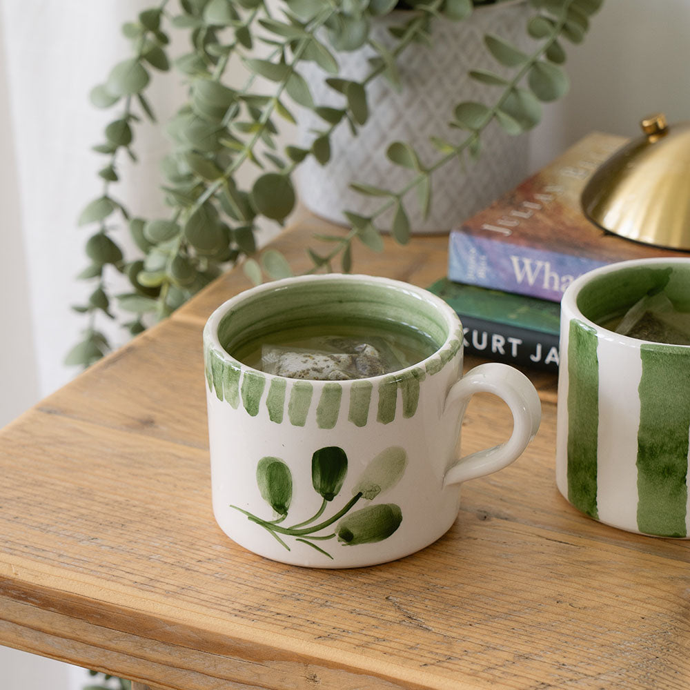 Hand-painted Selbrae House ceramic mug with green accents on a wooden table next to books and a plant