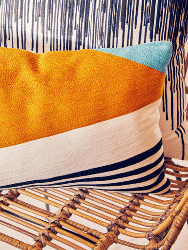 Orsina Embroidered Cushion with geometric patterns in orange, white, and blue on a woven chair