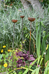 Three rustic metal Rain Catcher garden ornaments standing amid vibrant flowers and greenery