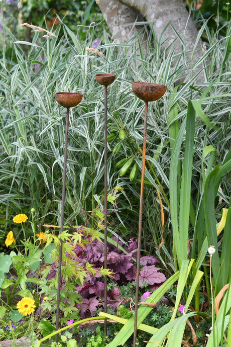 Three rustic metal Rain Catcher garden ornaments standing amid vibrant flowers and greenery