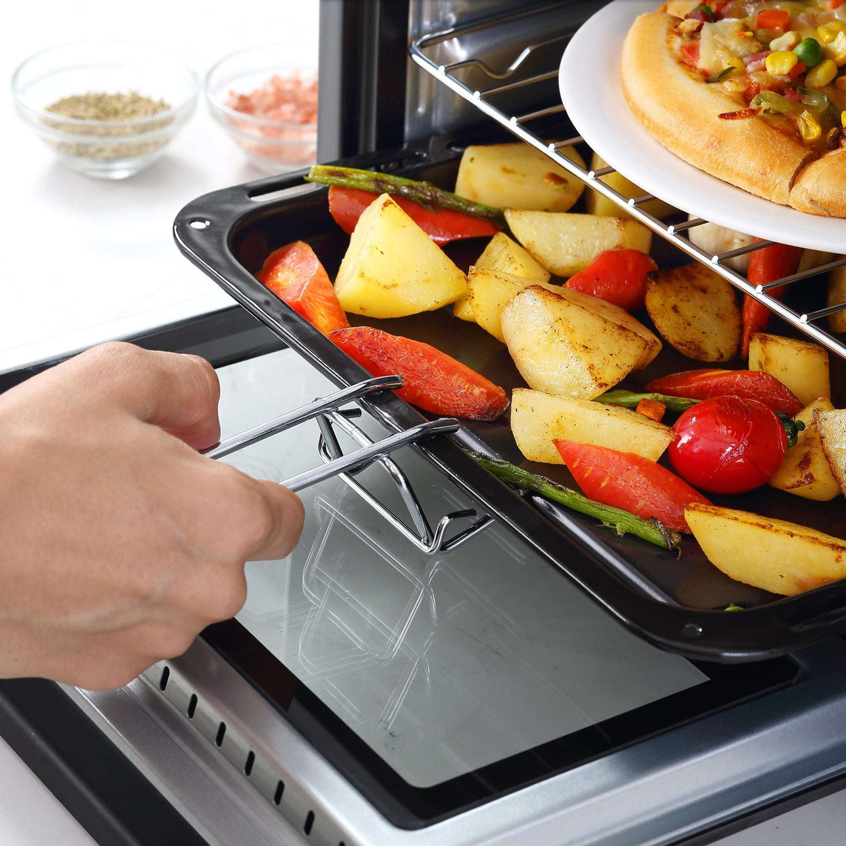 Hand removing roasted vegetables from a countertop oven with a dish of food above