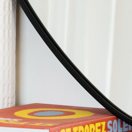 Jobu Home Pebble Mirror with a sleek black frame, hanging beside a decorative book for stylish decor