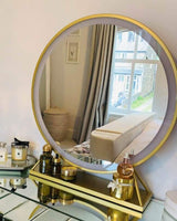 Gold Hollywood mirror reflecting a stylish room with decorative beauty products on a glass table