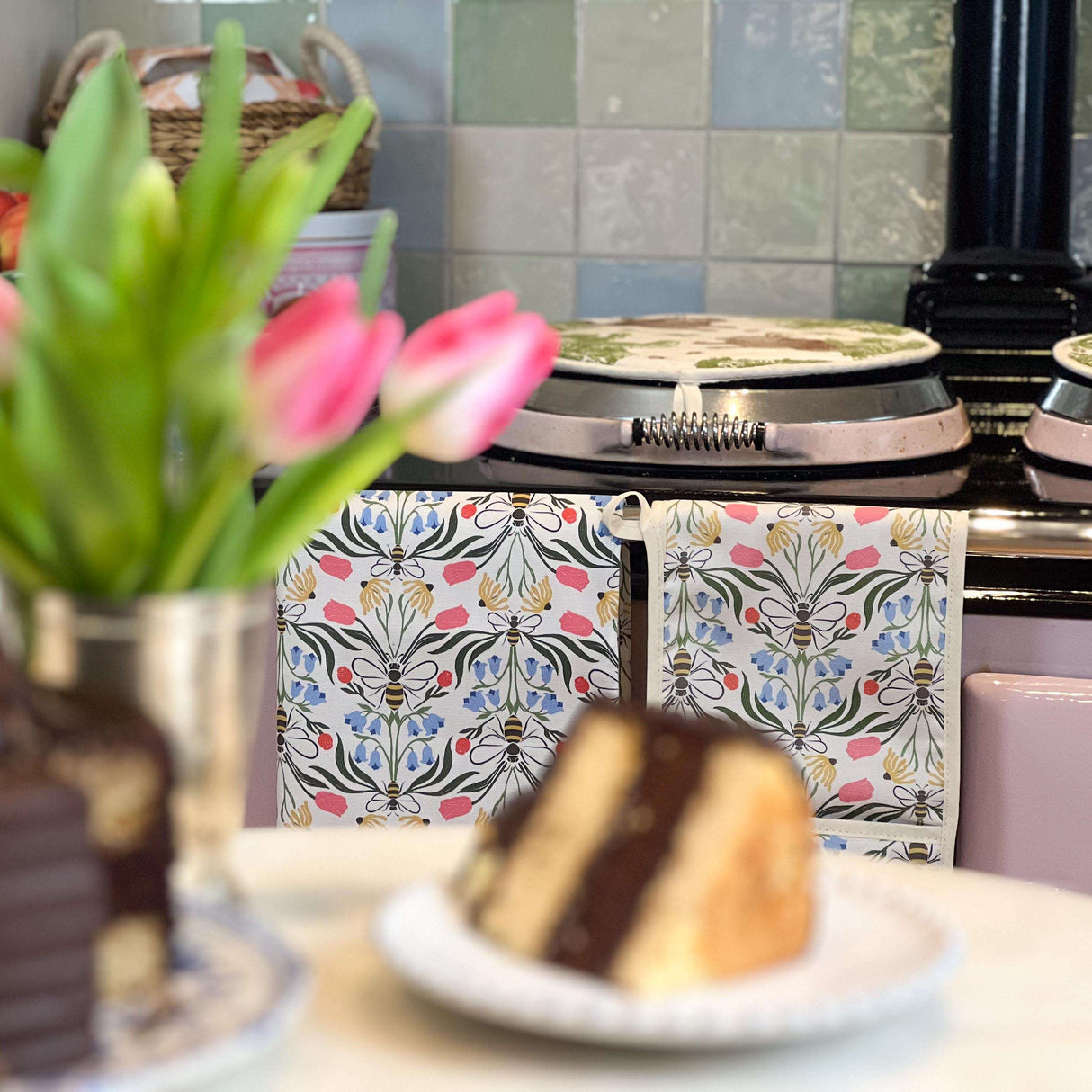 Floral Avalon Home UK Oven Gloves hanging near an Aga cooker with a slice of cake and fresh flowers
