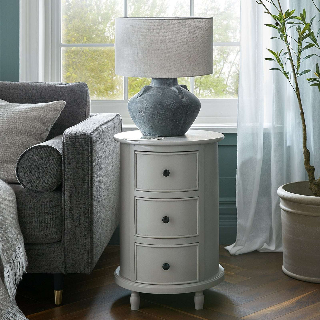 Shades 4 Seasons Round Side Table with three drawers and a stylish lamp in a cosy living room setting