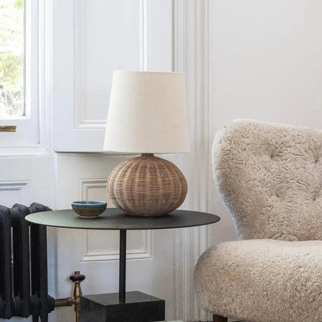 Nisu Home Table Lamp with a woven base and a beige fabric shade, placed beside a cozy armchair