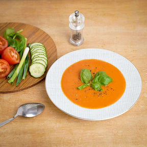 Elegant white soup bowl filled with tomato soup, garnished with basil, on a wooden table with fresh vegetables