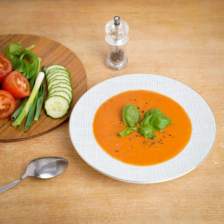 Elegant white soup bowl filled with tomato soup, garnished with basil, on a wooden table with fresh vegetables