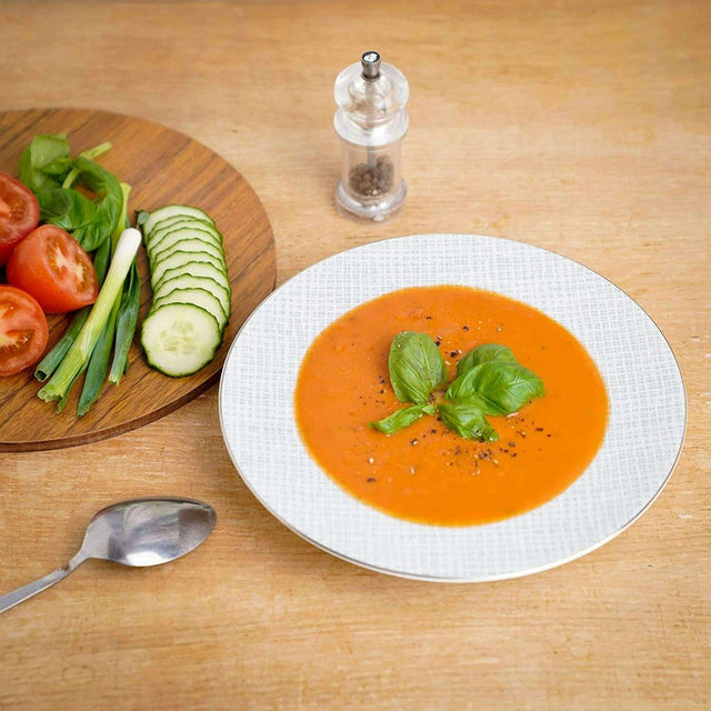 Elegant white soup bowl filled with tomato soup, garnished with basil, on a wooden table with fresh vegetables